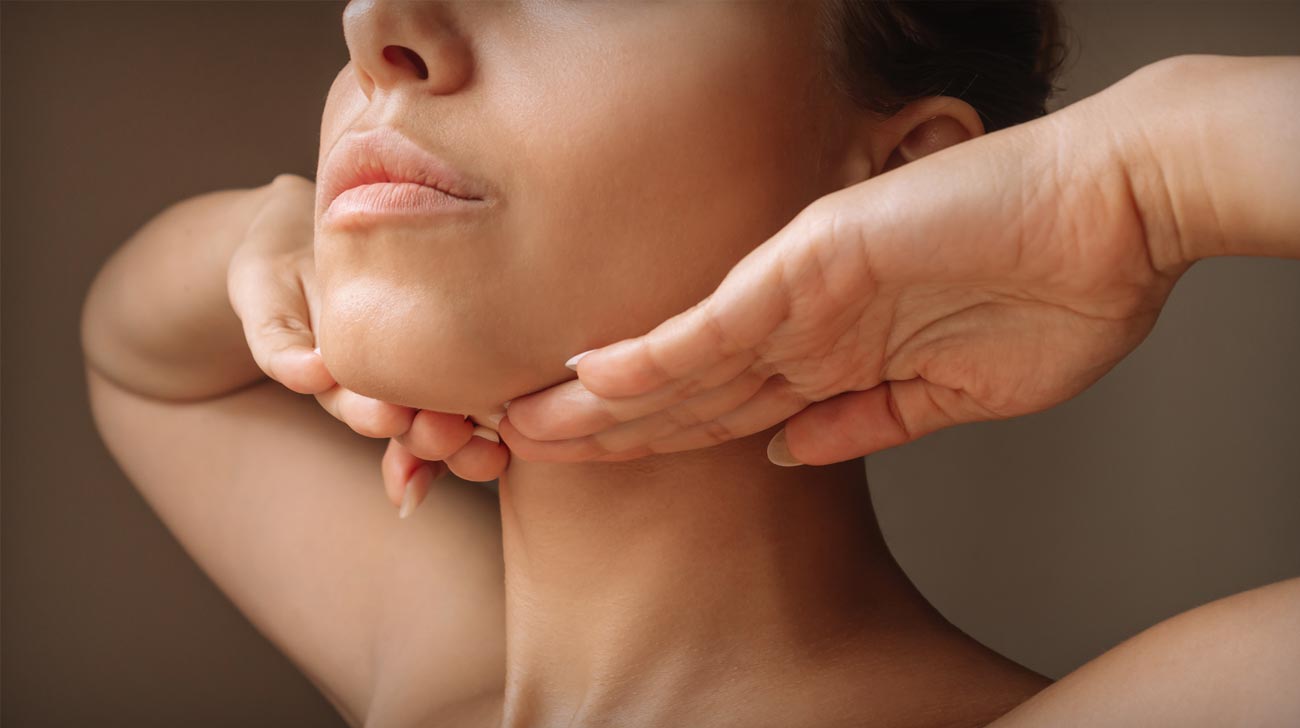 Cropped shot of young caucasian woman touching under the chin with hands massaging her face on dark brown background. Rejuvenation, facelift, facefitness.