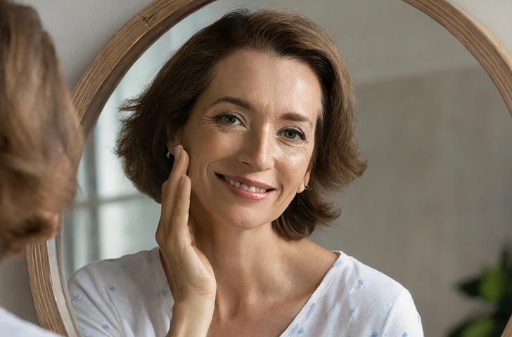 Happy 50s middle aged woman model touching face skin looking in mirror reflection. 