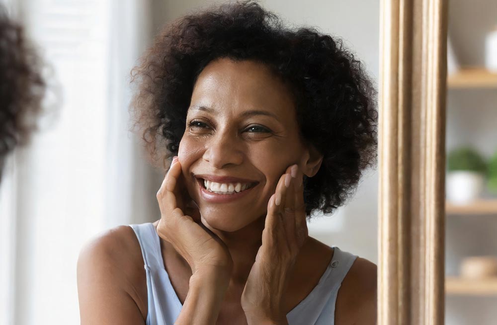 Happy 50s middle aged woman model touching face skin looking in