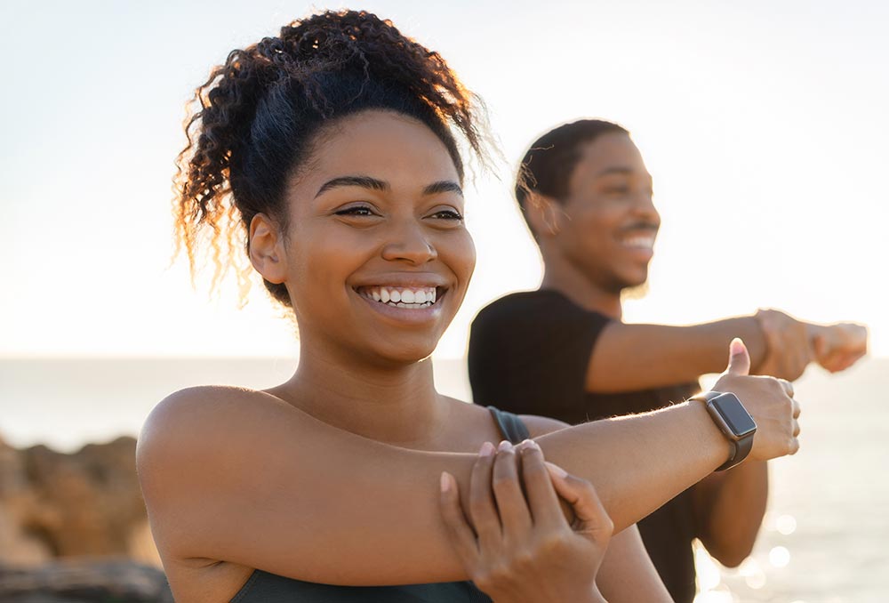 African American woman and man are stretching their arms in front of a beautiful sunset view over the ocean.