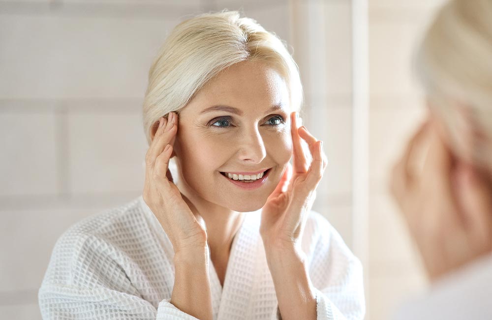 Gorgeous mid age adult 50 years old blonde woman standing in bathroom after shower touching face