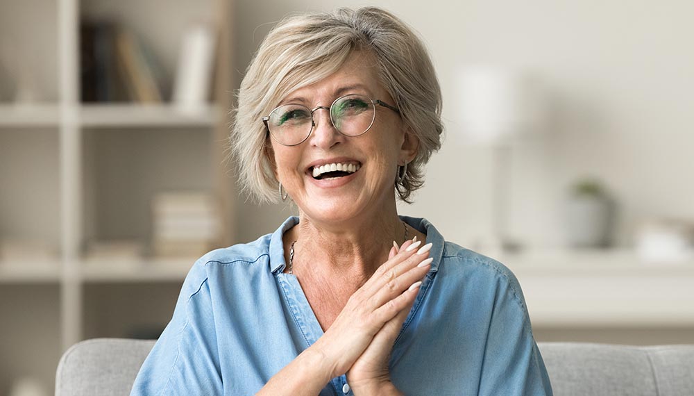 Cheerful pretty older woman in elegant glasses sitting on cozy home couch