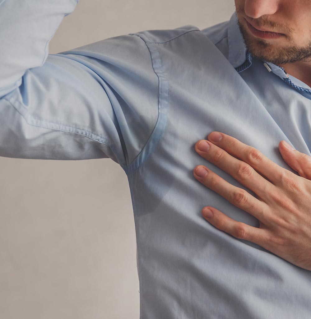 Man with hyperhidrosis sweating very badly under armpit in blue shirt, on grey.